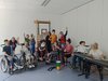 fünf Schüler/innen in einem Rollstuhl winken in die Kamera, daneben steht ein Keyboard und im Raum mehrere Musiker/innen