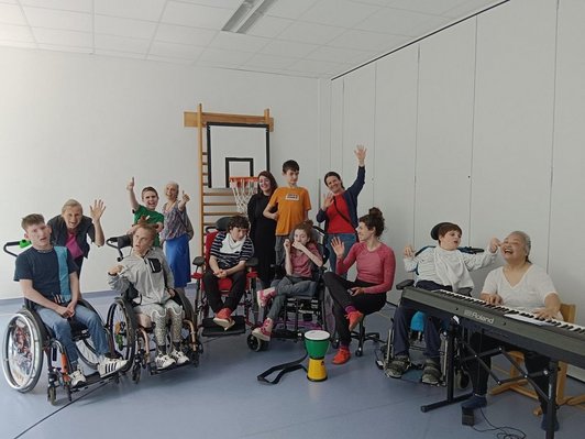 fünf Schüler/innen in einem Rollstuhl winken in die Kamera, daneben steht ein Keyboard und im Raum mehrere Musiker/innen