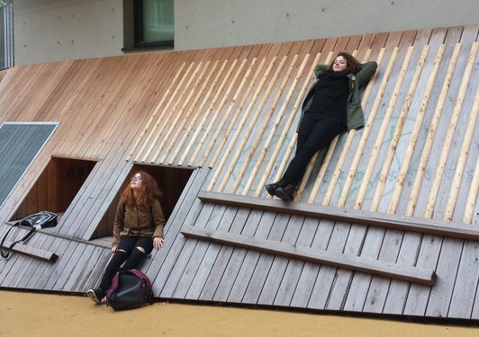 Foto von zwei Schülerinnen vor einer schrägen Holzfassade, eine sitzt in einer Ausnehmung, eine lehnt stehend