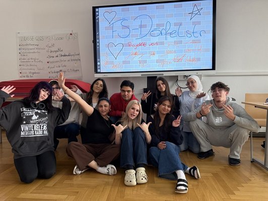 Gruppenbild von Schüler:innen vor einem Monitor mit eingeblendeter Mauer mit Schriftzug FS- Dörfelstraße und Unterschriften.
