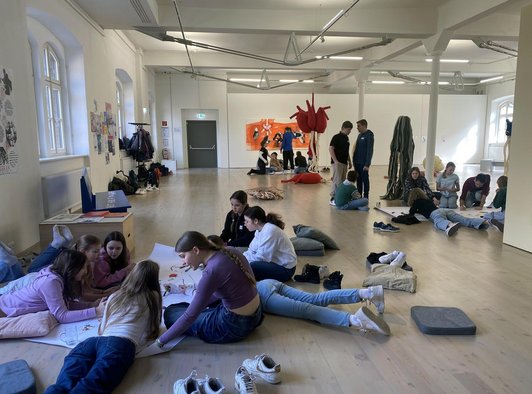 Schüler/innen liegen, sitzen und stehen in Gruppen und zeichnen auf großen Papierbögen