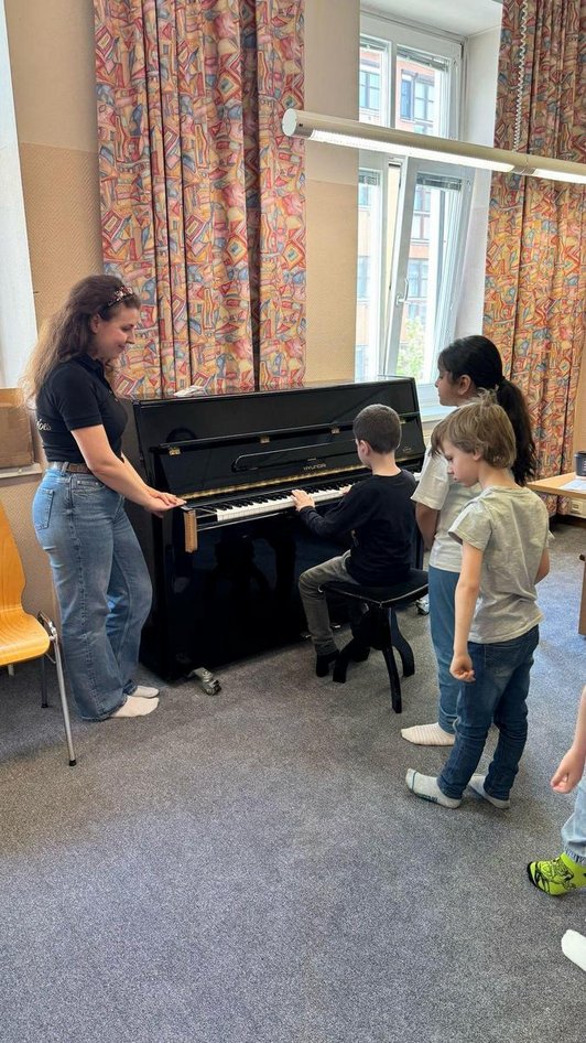 Kind spielt Klavier, weitere stehen in Reih dahinter und Frau lächend danaben