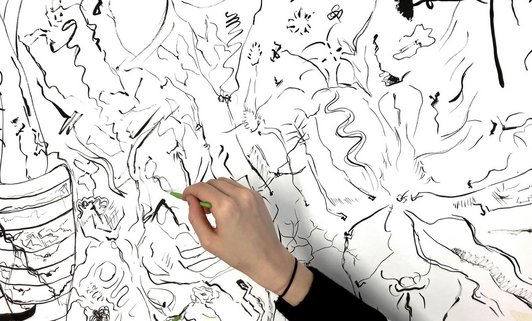 Foto von einer weissen Wand mit schwarzen Zeichnungen im Vordergrund eine Hand mit einem grünen Stift zeichnend.