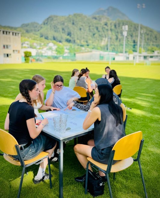 10 Schülerinnen sitzen an zwei quadratischen Tischen in Wiese
