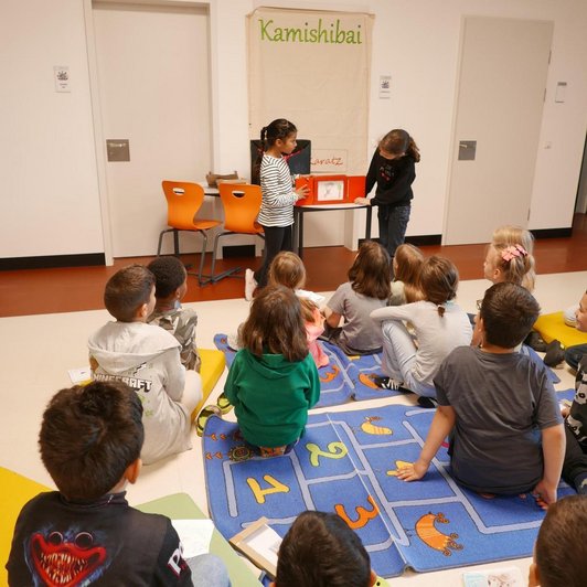 zwei Kinder präsentieren vor auf Boden sitzender Schulklasse