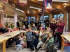 Foto von Kindern mit selbstgemachten aus recyceltem Material Musikblasintstumenten. Sie knien im Vordergrund um sie herum sitzen ältere Menschen und hören zu. Dahinter steht noch rine Fastnachtmusikgruppe mit Blasinstrumenten.