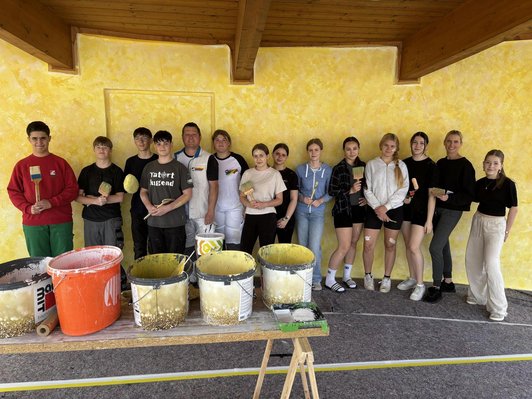 Fot ovon Schüler:innen vor einer gelben Wand mit Pinseln in der Hand. Im Vordergrung gelbe Farbkübel.