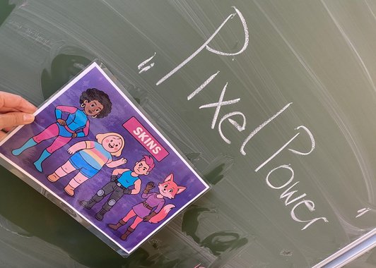 Auf einer Schultafel steht in Kreide geschrieben "Pixel Power". Davor hält ein Kind einen Ausdruck eines Comics mit dem Begriff "Skins"