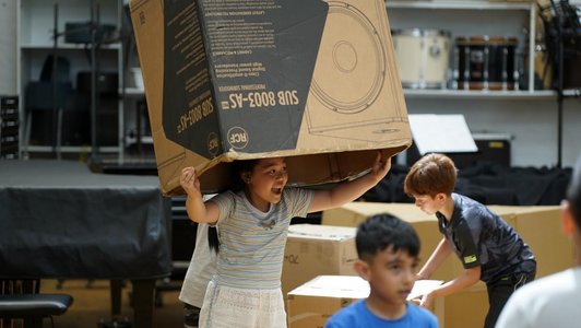 Ein Mädchen hebt eine große Kartonschachtel in die Luft, zwei weitere Kinder bewegen sich in weiteren Schachteln.
