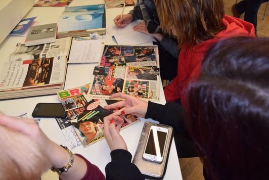 Foto von vier SchülerInnen von hinten, die rund um eine Tisch mit Zeitungsausschnitten, aufgeschlagenen Büchern und Mobiltelefonen sitzen, ein macht Notizen.