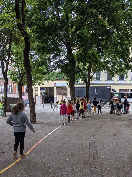 Foto von Schüler/innen, die auf eine bunten Linie unter Bäumen  im öffentlichen Raum balancieren