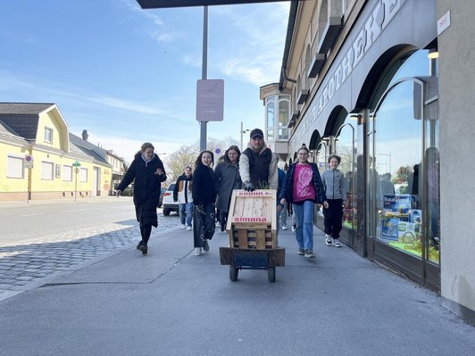 Fünf Jugendliche in einer Straße mit Geschäften schieben einen selbstgebauten Würfel mit der Aufschrift "simona - simona"
