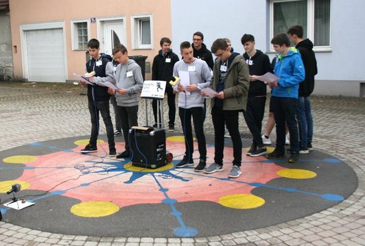 Foto von 12 Schülern, die mit Textheften und zwei Mikrofonen im öffentlichen Raum performen