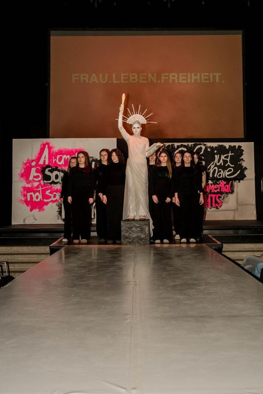 Foto von Schülerinnen in schwarz gekleidet auf einem Laufsteg. Eine Person als Freiheitsstatue in weiß in der Mitte.
