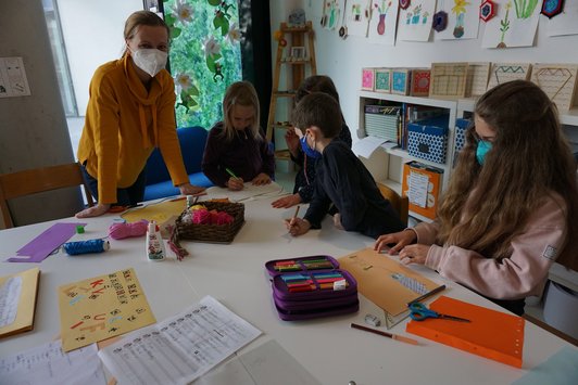 'Foto der Künstlerin und von vier Schüler/innen, die Buchseiten gestalten auf dem Tisch liegen Schreib- und Bastelmaterialien.'