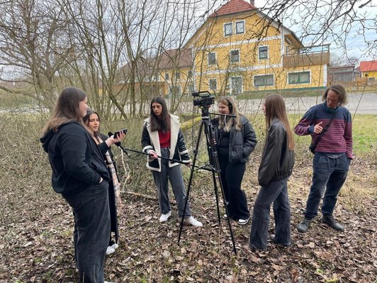 Schülerinnen und Mann stehen in Dorfumgebung um Kamera auf Stativ