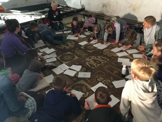 Foto von Schüler/innen, die mit drei Erwachsenen im Kreis auf Pölstern und Teppichfliesen sitzen und zeichnen