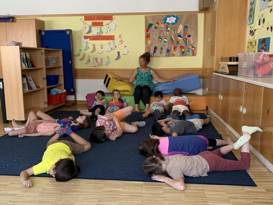 Kinder liegen in Klassenzimmer auf Teppich am Boden, Frau sitzt im Hintergrund