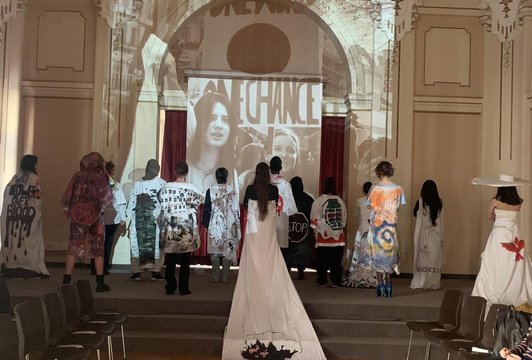 Schüler/innen stehen mit dem Rücken zum Bild und haben weiße selbstgenähte Kleider, die bedruckt sind. Darüber liegt eine Projektion "Give Peace a Chance".