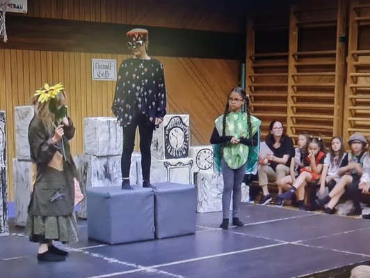 drei Schülerinnen in Kostümen auf Bühne, Kinder-Publikum im Hintergrund