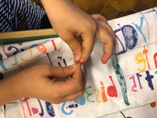 Foto von den Händen eines Kindes beim Verknoten eines Fadens auf der Rückseite eines Stücks Stoff mit gemalten bunten Buchstaben