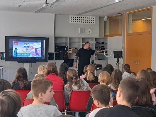 Schüler/innen sitzen in einer Reihe und schauen auf einen Bildschirm, vorne steht ein Referent und erklärt das mitgebrachte Equipment.