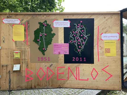 Ausstellungswand im Freien mit Plakaten und dem großen Schriftzug "BODENLOS" in pink