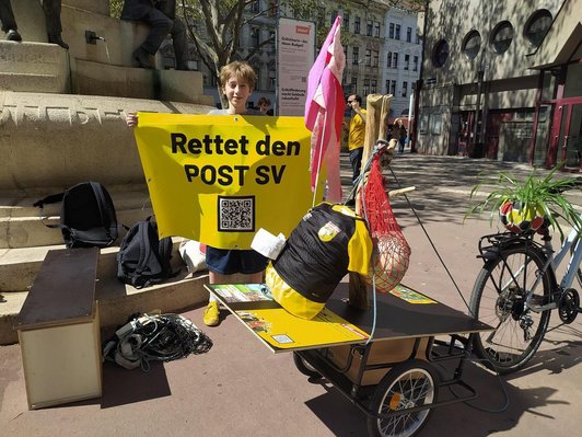 Foto voneinem Schüler mit einem mobilen Infotisch über einen Fahrradhänger hält ein gelbes Plakat mit dem Schriftzug "Rettet den POST SV"