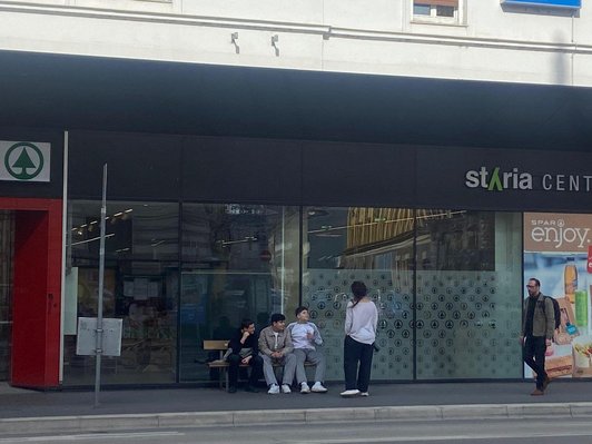 drei Schüler sitzen auf Bank vor Supermarkt, eine Schülerin steht daneben