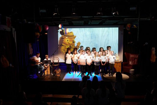 18 Kinder stehen vor Projektion eines Bären auf einer Bühne, ein Bub und ein Mann musizieren daneben