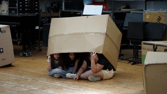 drei Mädchen blicken unterm Rand einer Kartonschachtel hervor, die zwei von ihnen anheben
