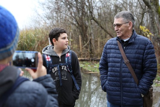 Foto eines Schülers im Gespräch mit einem Senioren, die in der Natur von einem zweiten Schüler mit dem Handy gefilmt werden 