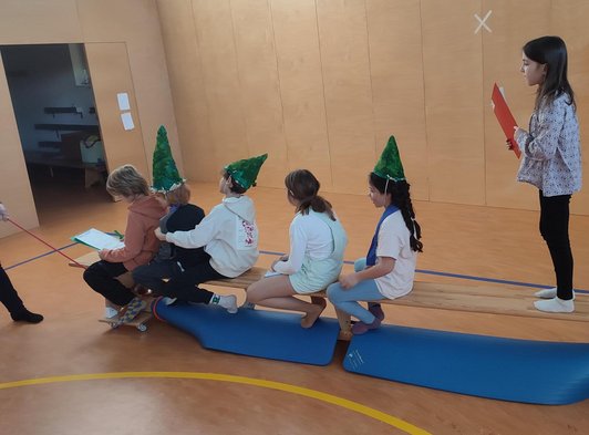 sechs Schüler/innen, teils mit gründen Hüten sitzen und stehen hintereinander auf Turnbank