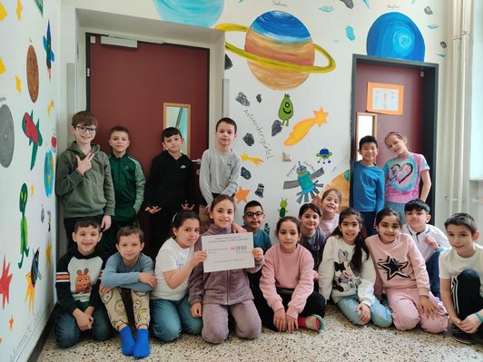 Schulklasse von Volksschüler/innen sitzen vor einer Wandmalerei eines Weltalls im Stiegenhaus der Schule.