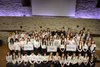 Foto von Schüler:innen auf einerbÜhne von schräg oben, alle mit eißen Oberteilen und schwarzer Hose . Drei haben Schilder in der Hand.