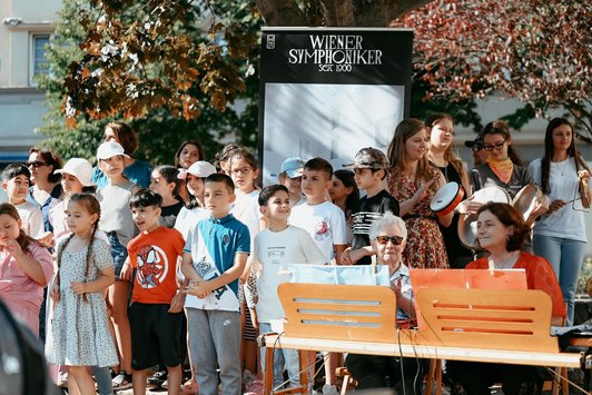 Eine Volkschulklasse steht hinter zwei Musiker/innen, die vor einem Notenbrett sitzen.