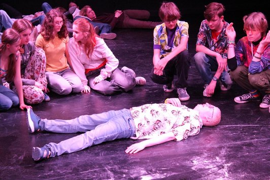 Schüler/innen hocken und knien auf Bühne um auf Rücken liegenden Mann