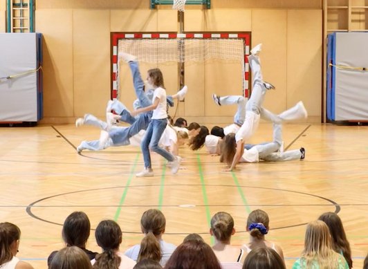 Mädchen in Jenas und weißen T-Shirts tanzen in Turnsaal, Publikum im Vordergrund