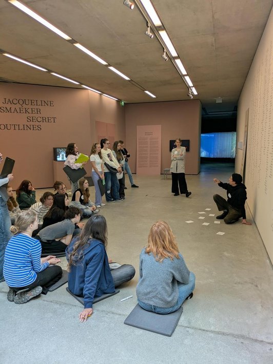 Schüler/innen sitzen und stehen in Ausstellungsraum, Mann erklärt