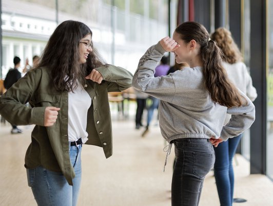 'Foto von zwei lächelnden Schülerinnen, die sich an den Ellbogen berüühren in Hintergrund sind weitere Schüler/innen zu sehen.'