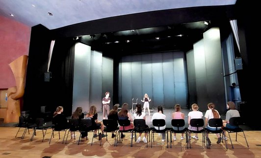 zwei Schüler/innen auf einer Bühne, 11 weitere in einer Sesselreihe davor