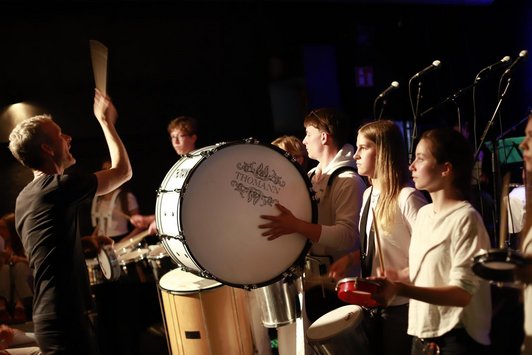 Dirigent mit erhobenem Stab vor Jugendlichen mit Percussion-Instrumenten 