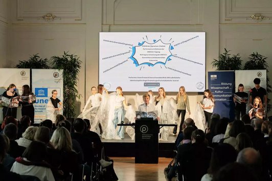 Bühne mit transparenten Foliern, ein Schüler sitzt im Vordergrund an einem Tisch mit Mikrophon und einem Unseco Papier in der Hand. Auf der Leinwand im Hintergrund steht auf verschiedene Sprachen "Poisie der (Sprachen-)Vielfalt.