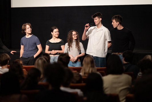 fünf Schüler/innen moderieren vor einer Schulklasse, die einem Kinosaal sitzen, den Ablauf. 
