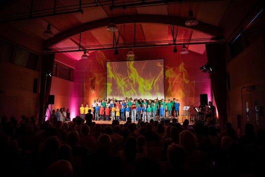 Bühne voller Kinder mit buntern Shirt und Projektion von Flammen