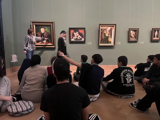 Schüler/innen sitzen am Boden in einem Kunstmuseum und diskutieren mit einer Kunstvermittlerin, die vor einem Bild steht.