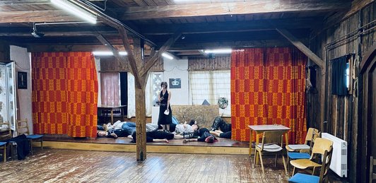 Auf einer kleinen Bühne steht eine Schüler/innen mit einem Handy in der Hand, neben ihr liegen mehrere Schüler/innen am Boden.