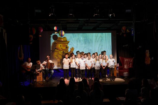 Eine Gruppe singender Kinder, eine kleines Ensemble, die begleiten und ein Bär, der projiziert wird.