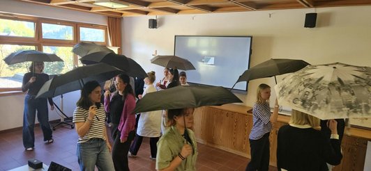 Foto von Schülerinnen mit Regenschirmen gehend in einem Zimmer mit Monitor.
