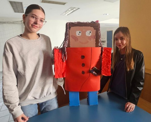 Foto von zwei Mädchen mit einer gebastelten Papierfigur ,mit orangem Hermd und blauer Hose in der Mitte.
